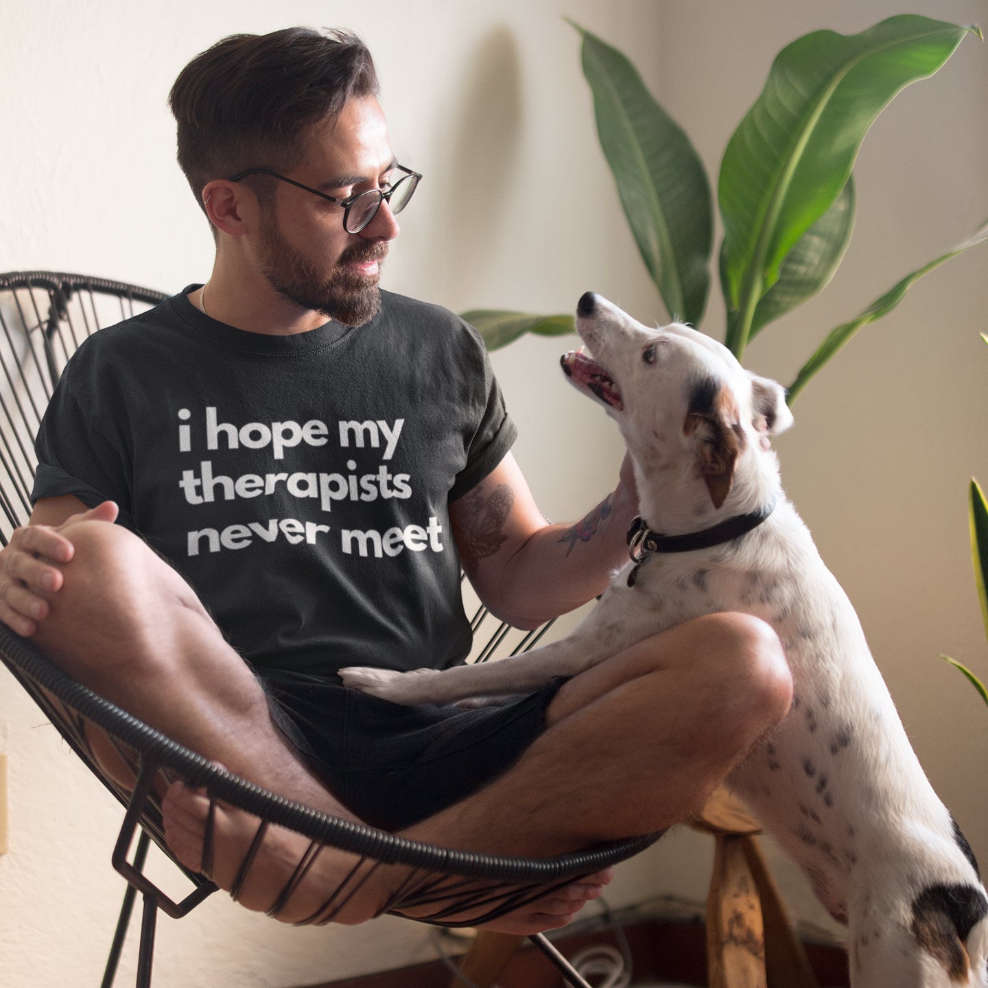 Man sitting on a chair with a dog, wearing a t-shirt with text, in a home setting.